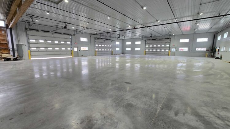 A wide, polished concrete floor inside a spacious, empty modern warehouse with multiple closed overhead garage doors and bright ceiling lights.