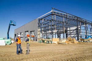 Two construction workers talking outside a large industrial steel-frame building under construction against a clear blue sky.